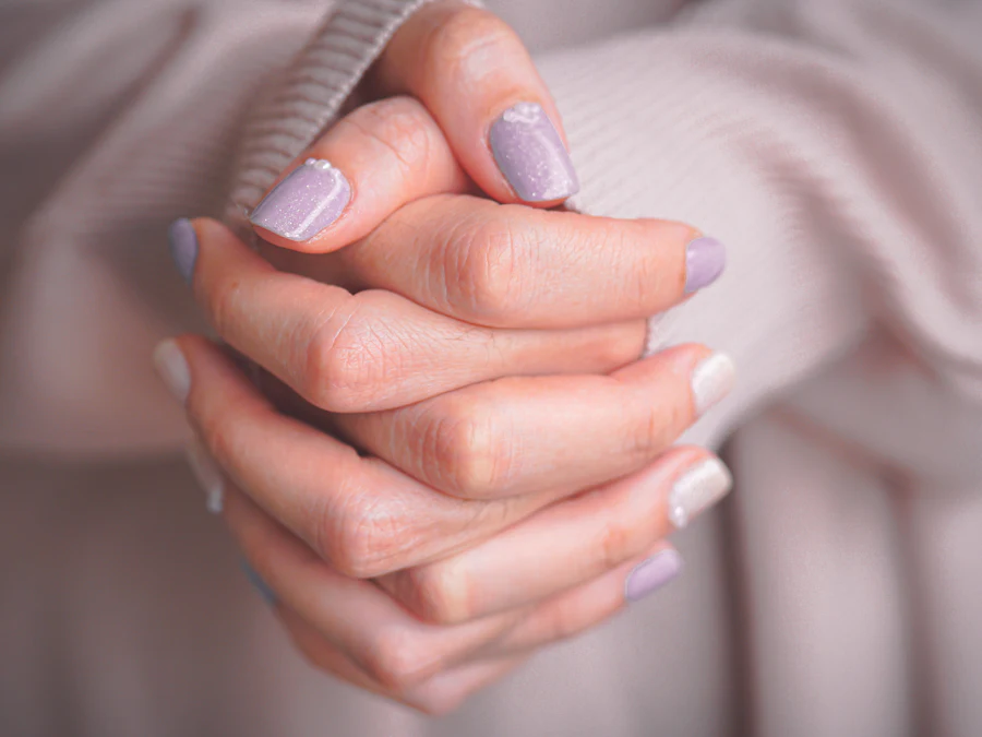 มือประสานกันในแขนเสื้อไหมพรมสีชมพูอ่อน เล็บเจลสีม่วงไลแลคมีกากเพชร สลับเล็บสีเงิน ติดมุกเม็ดเล็กที่โคนเล็บ